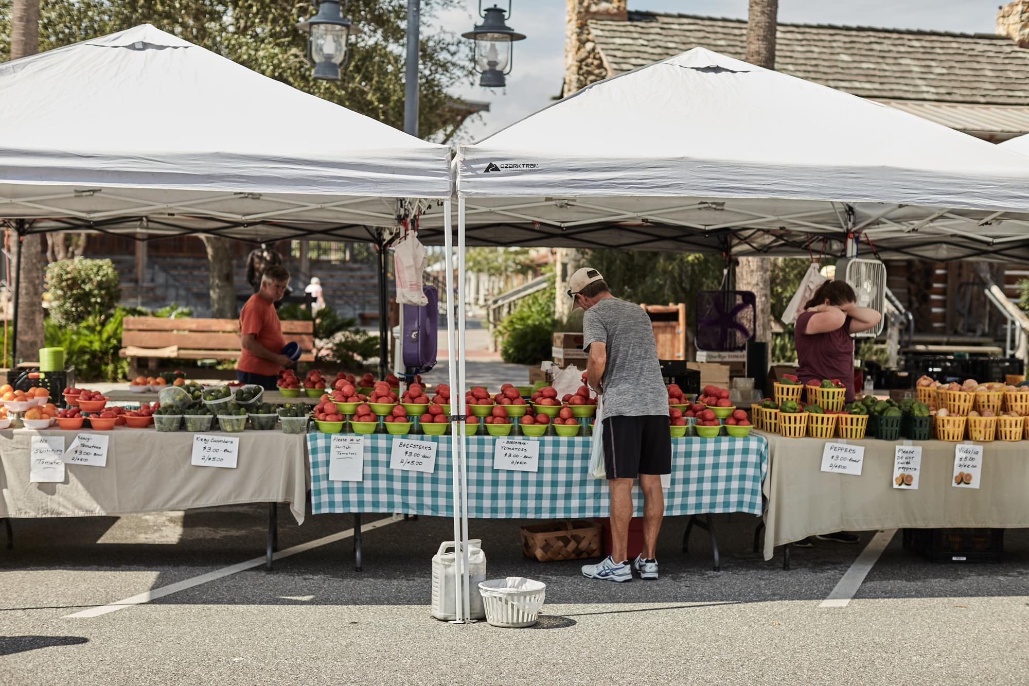 Shoppers browsing tomatoes and peppers under white tents at an outdoor farmers market